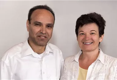 Two people are standing in front of a neutral background. Both are smiling friendly at the camera.