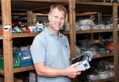 A man stands in a storage area holding a tool in his hand. In the background, shelves with various materials and supplies can be seen.