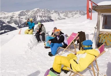Fünf Personen entspannen auf einem verschneiten Berg. Im Hintergrund sind schneebedeckte Berge und ein Holzhaus zu sehen.