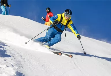 Ein Skifahrer in bunter Kleidung fährt die schneebedeckte Piste hinunter. Im Hintergrund sind weitere Skifahrer und ein klarer blauer Himmel zu sehen.