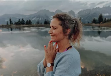 A woman stands at the edge of a calm lake and smiles. In the background, mountains and a cloudy sky are visible.