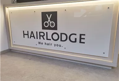 Ein modern gestaltetes Schild mit dem Namen „HAIRLODGE“. Darunter steht der Slogan „We hair you.“