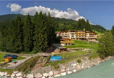 Eine malerische Landschaft mit einem Hotel am Fluss und grünen Wiesen. Im Hintergrund erheben sich majestätische Berge unter einem blauen Himmel.
