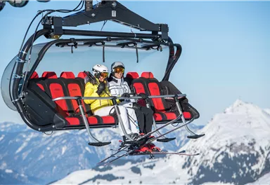 Eine Seilbahn mit zwei Skifahrern, die in den Bergen sitzen. Im Hintergrund sind schneebedeckte Gipfel und ein klarer blauer Himmel zu sehen.