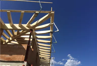 Ein Dachstuhl aus Holz, der im Bau ist, steht vor einem klaren blauen Himmel. Im Hintergrund sind Berge und einige Wolken sichtbar.