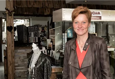 A woman is standing in a clothing store and smiling at the camera. In the background, various garments and accessories can be seen.