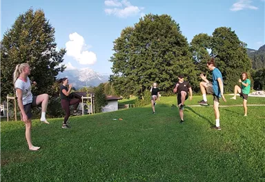 A group of people is exercising on a meadow. Trees and mountains are visible in the background.