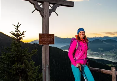 Eine Wanderin steht an einem Holzkreuz auf einem Berg. Im Hintergrund sind die Berge und ein klarer Himmel zu sehen.
