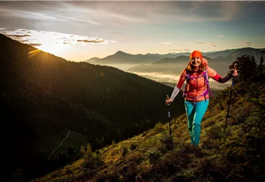 Eine Frau wandert in den Bergen bei Sonnenaufgang. Die Landschaft ist grün und malerisch mit einem klaren Himmel im Hintergrund.
