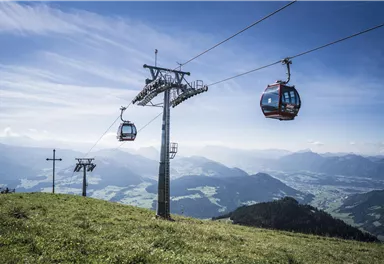 Eine Seilbahn fährt über eine grüne Wiese mit Bergen im Hintergrund. Der Himmel ist klar und blau.