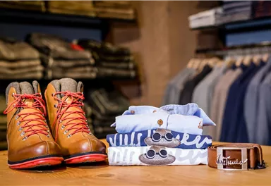 Ein Paar braune Wanderstiefel mit roten Schnürsenkeln steht auf einem Holztisch. Daneben liegen Kleidung und ein Gürtel in ansprechenden Farben.