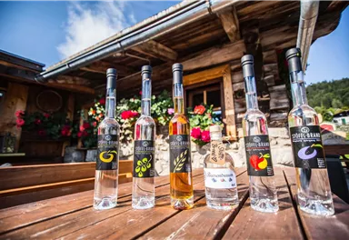 A row of bottles with various schnapps is standing on a wooden table. In the background, you can see a traditional alpine hut with colorful flowers.