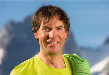 Ein sportlicher Mann mit kurzen Haaren lächelt in die Kamera. Er trägt ein grünes Shirt und hält ein Kletterseil in der Hand, während die Berge im Hintergrund sichtbar sind.