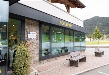 A modern store named "SCHUHHAUS" in a nature-friendly environment. Benches stand in front of the store and there is limited space for greenery.