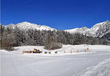 A snowy landscape with high mountains in the background. Some shoveling and seating areas are visible, surrounded by snow-covered trees.