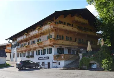Ein charmantes Gebäude im bayerischen Stil mit großen Fenstern und Balkonen. Vor dem Haus stehen Autos und Sonnenschirme auf der Terrasse.