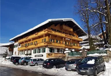Ein traditionelles alpenländisches Gebäude mit einem Holzbalcon und grünen Fensterläden. Die Umgebung ist schneebedeckt und es stehen mehrere Autos auf der Straße.