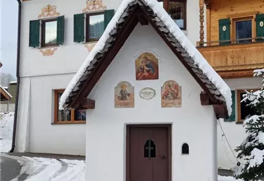 A small chapel with a snow-covered roof and intricate frescoes. In the background, traditional wooden houses can be seen.