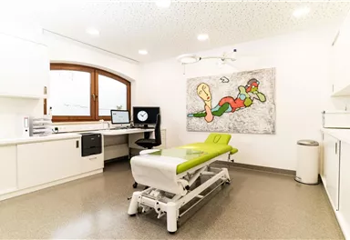 Ein modernes Behandlungszimmer mit einem grünen Behandlungstisch und einem bunten Kunstwerk an der Wand. Der Raum ist hell und aufgeräumt, mit einem Computer und medizinischen Geräten.