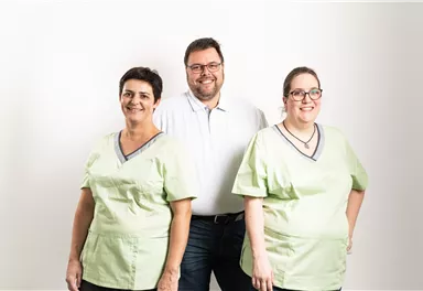 Ein sympathisches Team von drei Personen, zwei in grünen Arbeitskleidung und eine Person in einem weißen Hemd. Sie stehen vor einem neutralen, hellen Hintergrund.