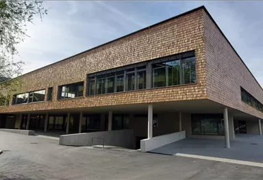 A modern building with wooden cladding and large windows. It is situated in a green environment and features a tidy architecture.