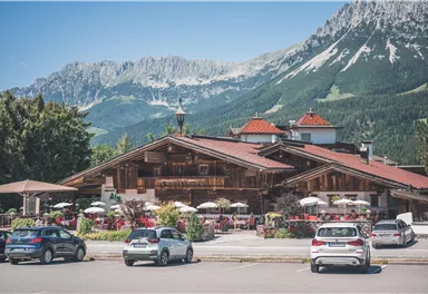 A traditional chalet in the mountains with blooming flowers and outdoor seating. In the background, majestic mountains and a clear blue sky are visible.