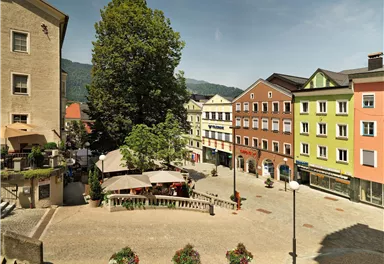 Eine lebendige Stadtansicht mit bunten Gebäuden und einem großen Baum. Tische im Freien laden zum Verweilen ein.
