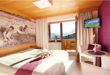 A cozy bedroom with a large bed and a beautiful view of the mountains. The walls are warmly decorated, and there is a television as well as a small table.