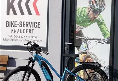 A blue e-bike is standing in front of a shop named "Bike-Service Knaubert". In the background, there is a large image of a cyclist.