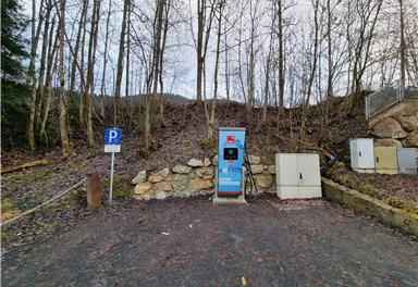 A charging station for electric cars is located in a rural area with trees in the background. Next to the station, there is a parking lot.