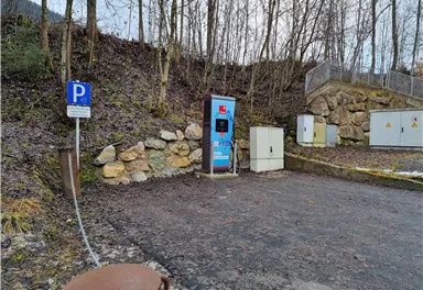 A charging station for electric cars stands in a parking lot in a wooded area. The ground is gravelly and there is a small rock wall in the background.