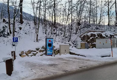 Ein Parkplatz in einer verschneiten Landschaft mit Bäumen. Im Vordergrund steht ein Parkautomaten.