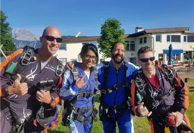 Vier Personen in Fallschirmanzügen stehen draußen und lächeln. Sie zeigen Daumen hoch an einem sonnigen Tag.