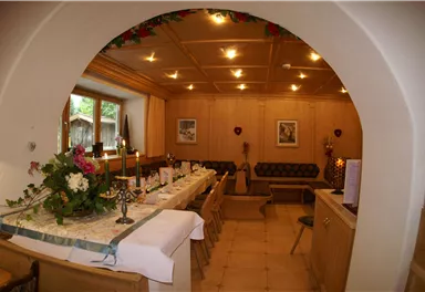 A cozy room with wooden paneling and a long table. Flowers and candles decorate the table, while in the background a comfortable seating area is visible.
