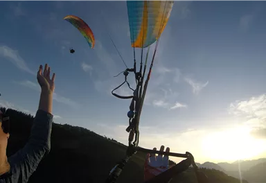 A paraglider is gliding over a picturesque landscape at sunset. A person is reaching out their hand and watching the paraglider.