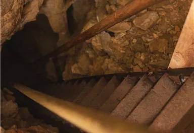 A steep metal staircase leads into a dark cave. The floor is covered with stones and earth.