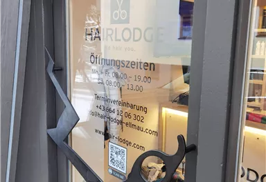 Ein Friseursalon mit einem modernen Eingang und einer Glasfront. Auf dem Fenster sind Öffnungszeiten und Kontaktinformationen angegeben.