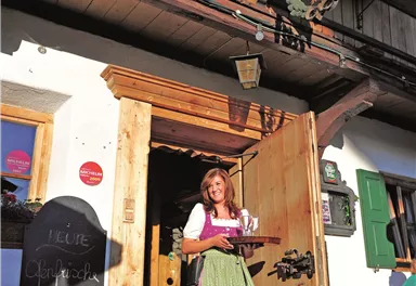 A woman in traditional attire stands in front of an inn, holding a tray with drinks. The facade of the house is adorned with flowers.