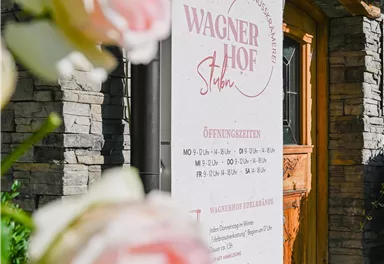 Ein Schild des Wagner Hofs mit Öffnungszeiten und Informationen. Im Vordergrund sind zarte rosa Rosen zu sehen.