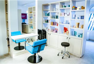 Ein modernes Friseurstudio mit hellen Wänden und Regalen voller Haarpflegeprodukte. In der Mitte steht ein Tisch mit einem blauen Stuhl und einem schwarzen Hocker.