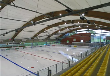 A modern ice rink with an empty ice field and a curved roof structure. The spectator stand is equipped with yellow seats.