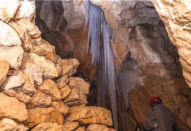 A cave with large rocks and stalagmites. A person with a red helmet is exploring the room.