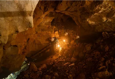 A gloomy cave chamber with rocks and a floor made of stones. Two people with helmets are holding flashlights and exploring the surroundings.