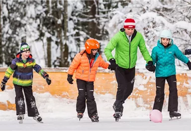 Eine fröhliche Gruppe von Kindern und einem Erwachsenen beim Schlittschuhlaufen auf einem Eisplatz im Schnee. Sie tragen bunte Winterkleidung und haben viel Spaß beim gemeinsamen Gleiten.