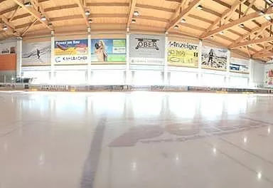 Eine Eishalle mit einer großen, leeren Eisfläche. Der Raum hat hohe Holzdecke und zahlreiche Werbetafeln an den Wänden.