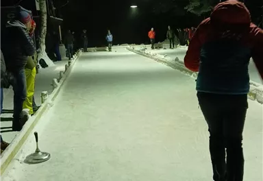 Eine verschneite Eisbahn bei Nacht mit beleuchteten Bereichen. Einige Personen sind beim Spielen aktiv.
