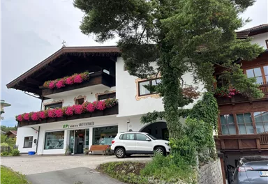 A traditional building with a balcony and blooming plants. In front of the house, there is a white car, and it is surrounded by trees.