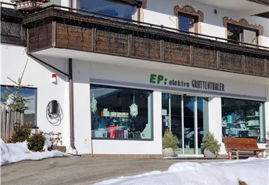An electronics store with large windows offers a view of various products. Snow lies in front of the store, and the surroundings are typical of a mountain landscape.