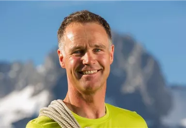 A smiling man is wearing a rope over his shoulder. In the background, mountains and a clear sky are visible.