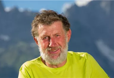 Ein lächelnder Mann mit grauen Haaren und Bart trägt ein leuchtend gelbes T-Shirt. Im Hintergrund sind schöne Berge zu sehen.